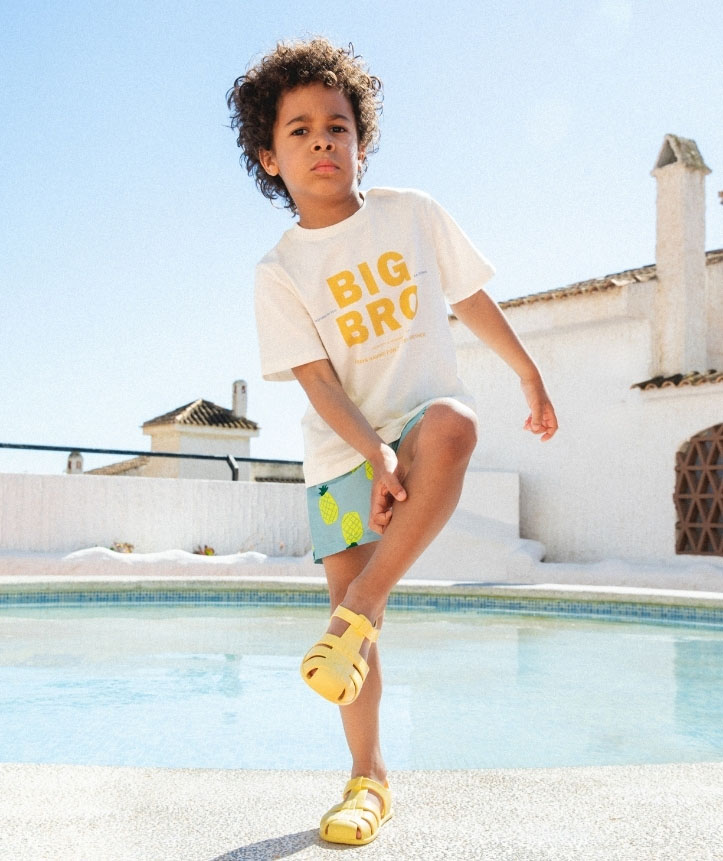 Criança com t-shirt branca, calções azuis com ananás e sandálias amarelas junto à piscina