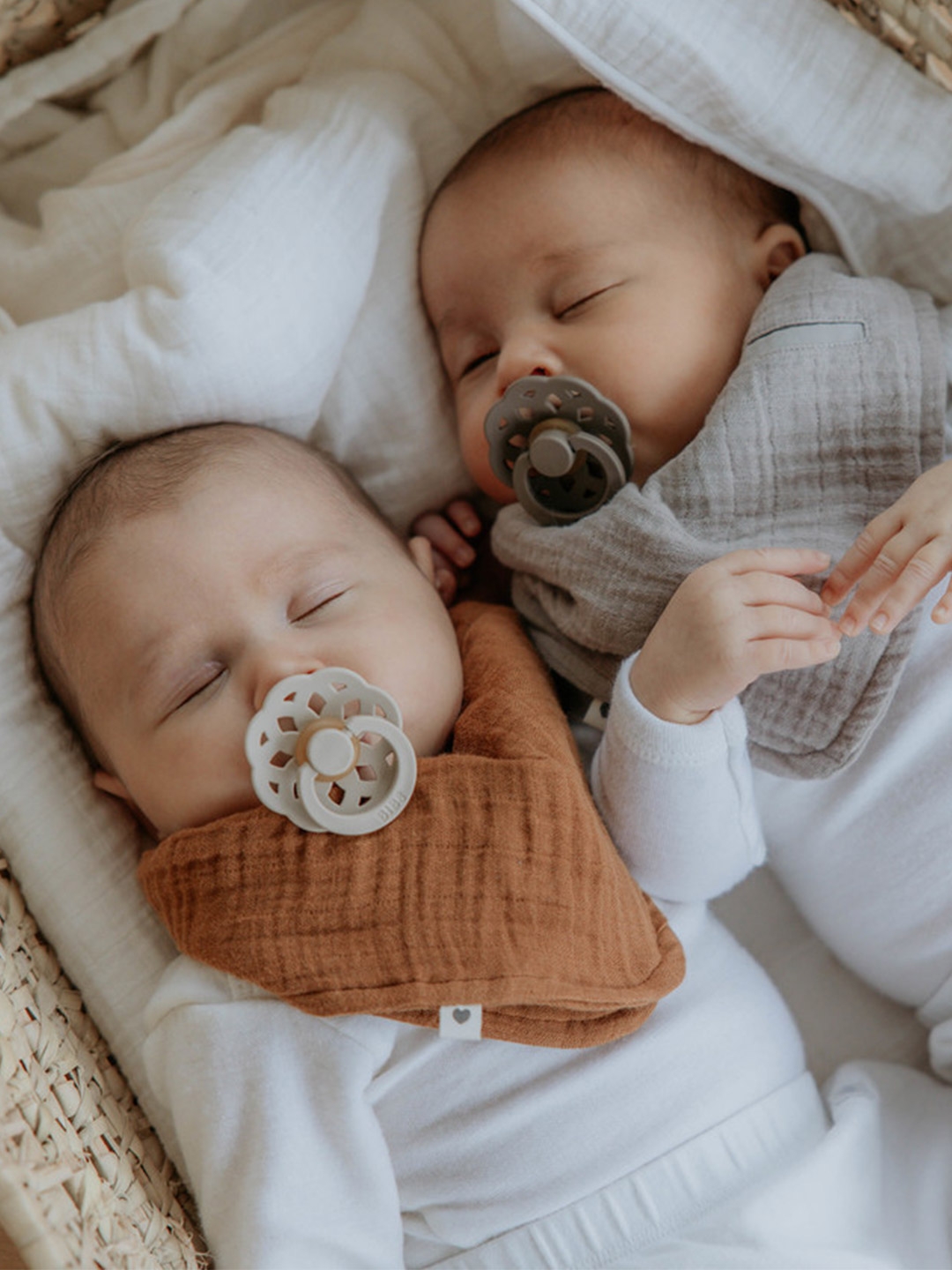 Dois bebés adormecidos com chupetas e babetes de tecido castanho e cinzento numa cama com lençóis brancos.