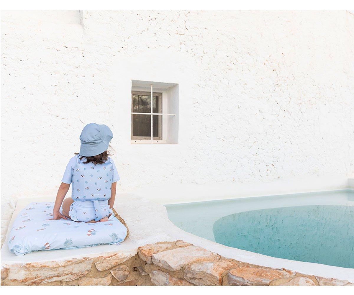 Criança de pijama azul e chapéu sentado numa almofada azul junto a piscina e parede branca