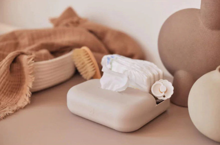 Caixa bege para lenços, toalha castanha e escova de madeira sobre superfície neutra