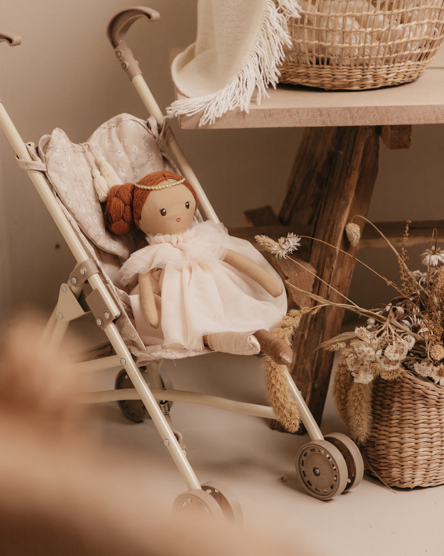 Carrinho de boneca com boneca de pano sentada e mesa com cesta de vime e flores secas