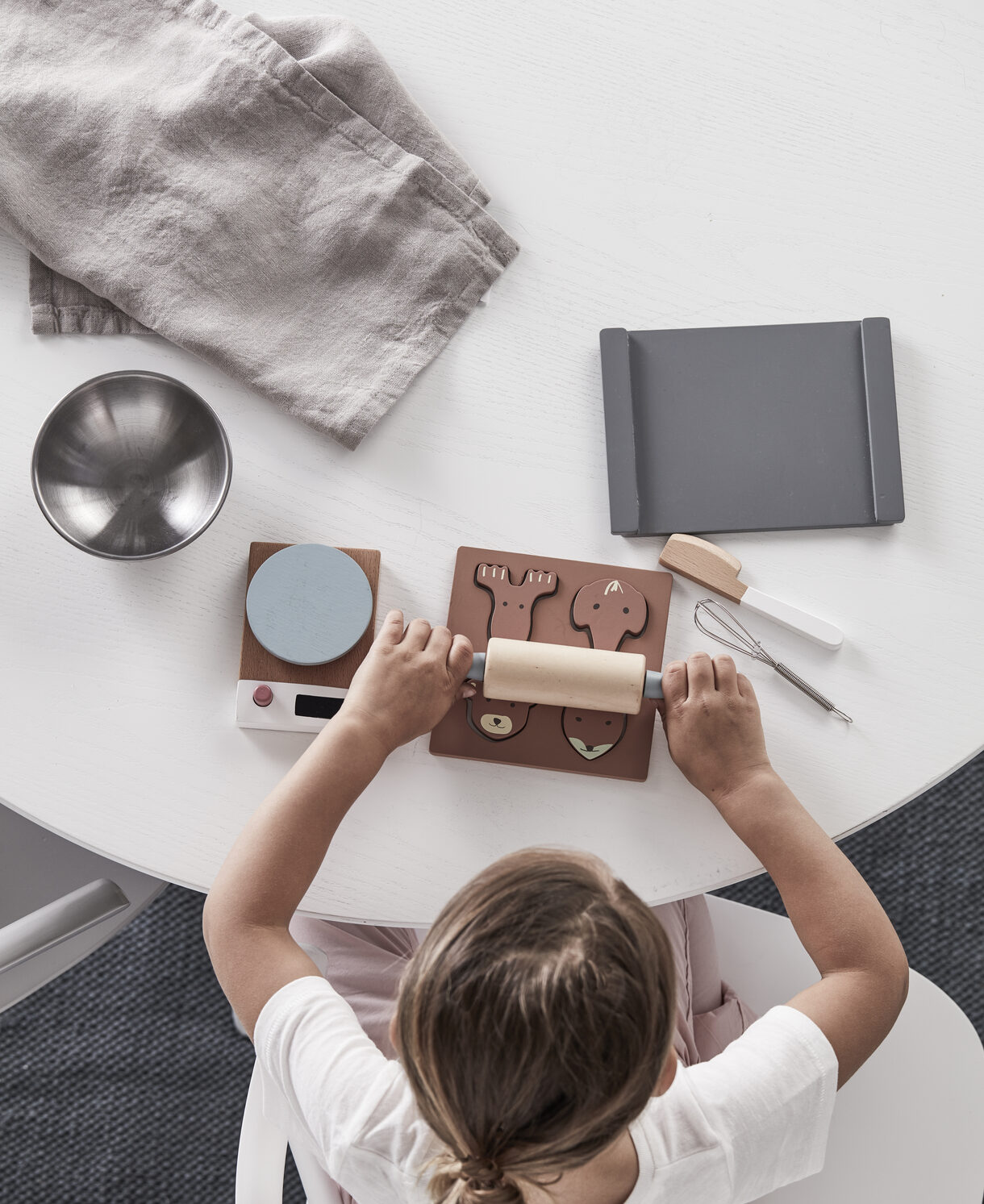 Criança a enrolar figuras de madeira com rolo sobre mesa com utensílios de cozinha e balança digital