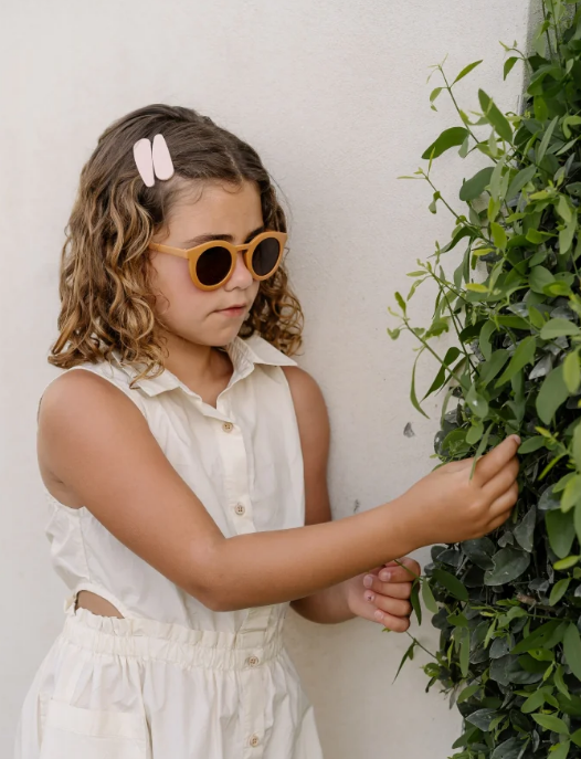 Menina com óculos de sol e vestido branco a tocar numa planta verde junto a uma parede branca