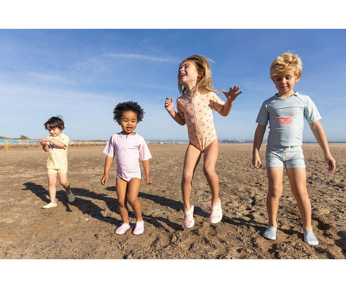 Quatro crianças a brincar na praia com fatos de banho variados, céu azul e areia ao redor