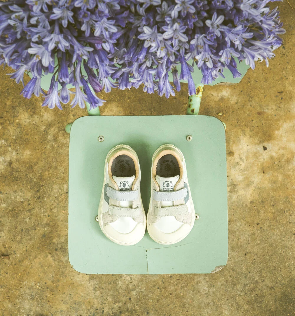 Sapatilhas infantis brancas com detalhes cinza em banco verde claro com flores roxas