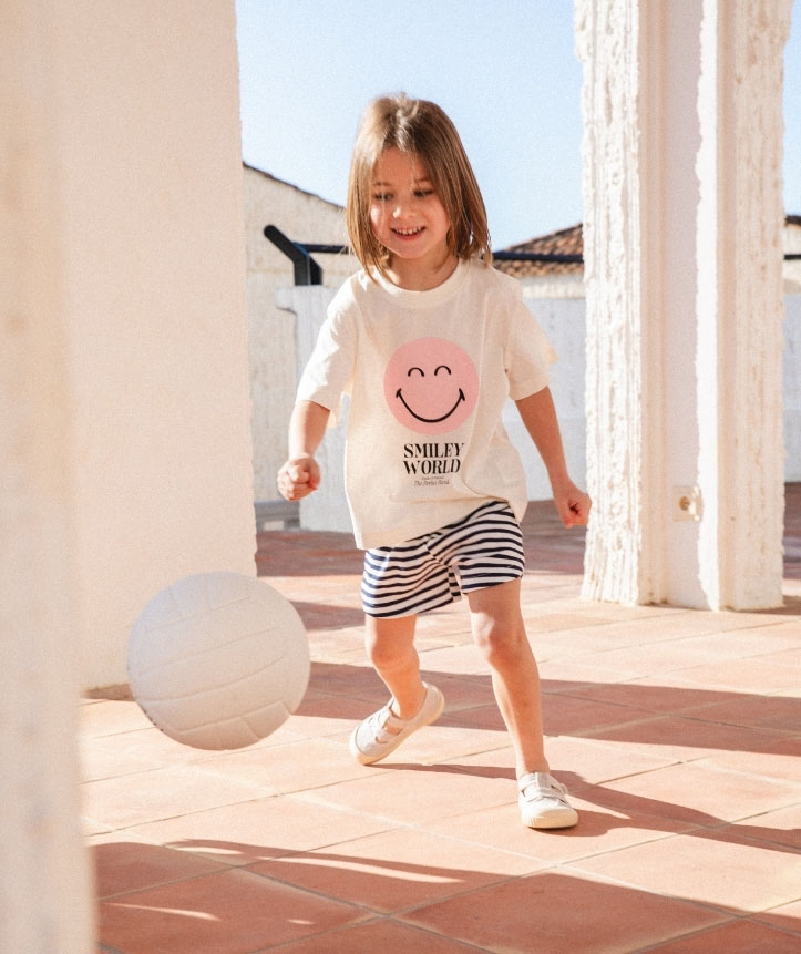 Criança a jogar com bola usando camisola branca com smiley e calções listrados