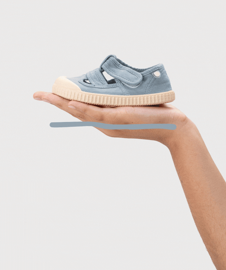 Sapato infantil azul claro em tecido com sola bege, estilo sandália com fecho de velcro, mão segurando sobre fundo branco