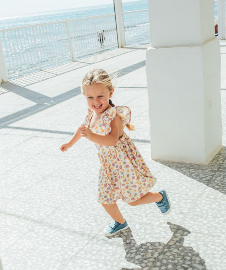 Menina a correr com vestido floral e ténis azuis num espaço ao ar livre junto ao mar