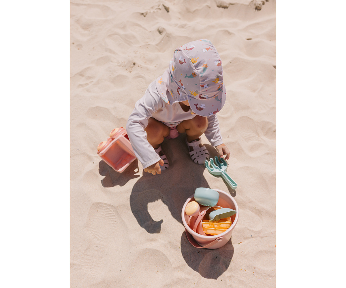 Criança a brincar com brinquedos de praia na areia, vestindo chapéu colorido e roupa branca.