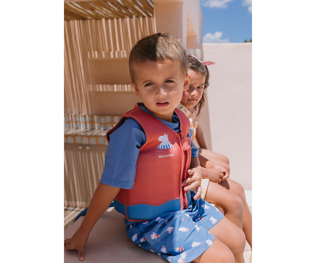 Menino com colete salva-vidas vermelho e calções azuis sentado ao lado de menina com fita cor-de-rosa no cabelo ao ar livre
