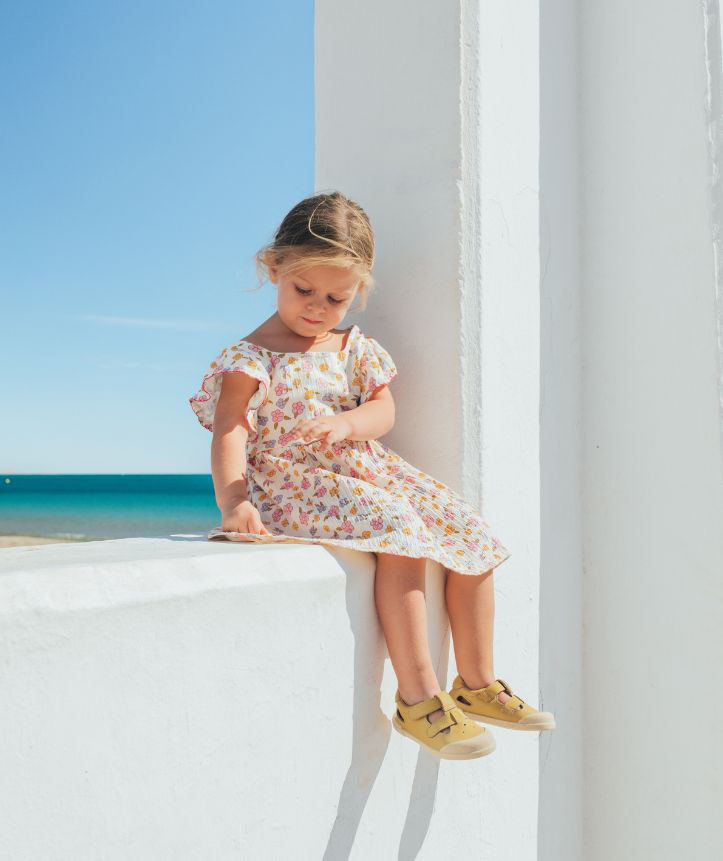 Criança com vestido floral e sapatos amarelos sentada numa parede branca com mar ao fundo
