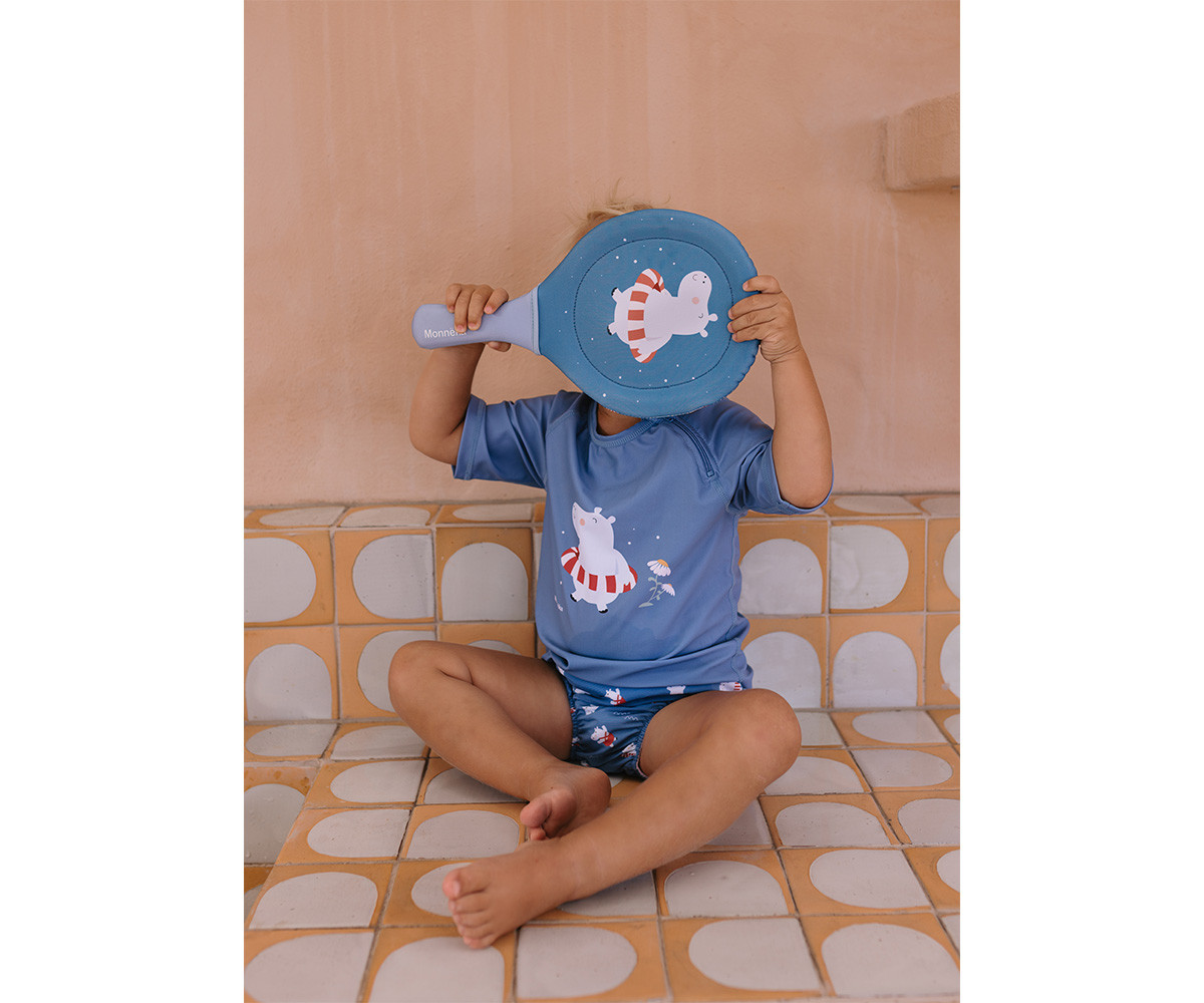 Criança a cobrir o rosto com raquete azul com estampa de urso polar sentada em azulejos bege e branco