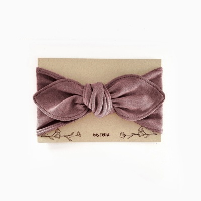 Faixa de cabelo em veludo lavanda com nó central em cartão bege com desenhos florais e marca.