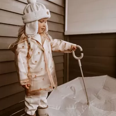 Menina com casaco bege segurando guarda-chuva branco aberto junto a parede de madeira
