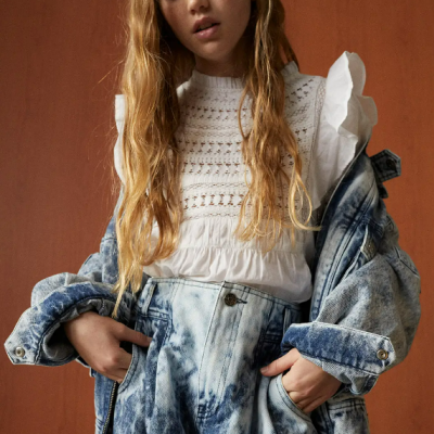 Menina com blusa branca de malha, jaqueta e calças de ganga azuis em fundo castanho