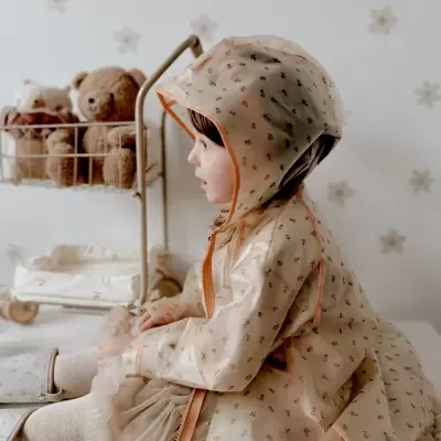 Criança com impermeável bege de padrão floral com capuz, sentada, com brinquedos atrás