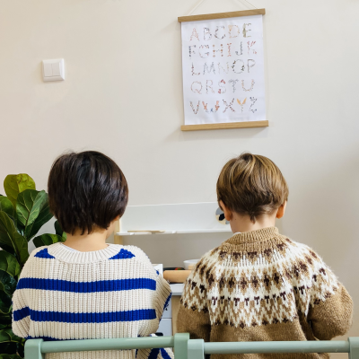 Duas crianças de costas sentadas em cadeiras verdes com suéteres de malha e parede decorada com quadro do alfabeto e borboletas.