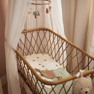 Berço de bebé em vime com almofada branca e mosquiteiro branco num quarto com ursinho de peluche e cortinas bege