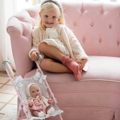 Criança sentada num sofá rosa ao lado de um carrinho de bebé de brinquedo com boneca.