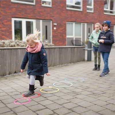 Criança saltando sobre argolas coloridas no chão de pedra com outros dois crianças e edifícios ao fundo