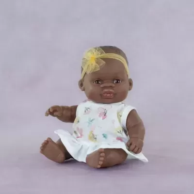 Boneca de bebé com vestido branco floral e fita amarela na cabeça sentada num fundo lilás.