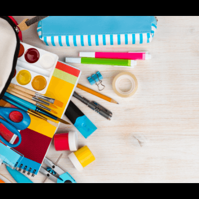 Material escolar colorido dentro e fora de uma mochila bege aberta sobre uma superfície de madeira clara.