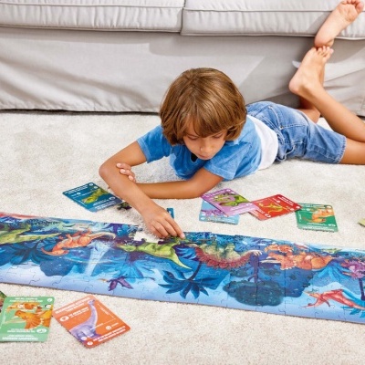 Criança a montar puzzle retangular de dinossauros coloridos no tapete com cartas de jogo ao redor