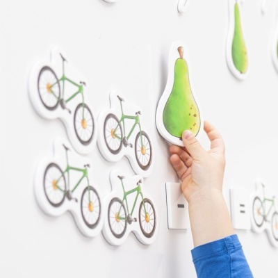 Imãs decorativos de bicicleta e pera numa superfície branca com mão de criança a segurar a pera