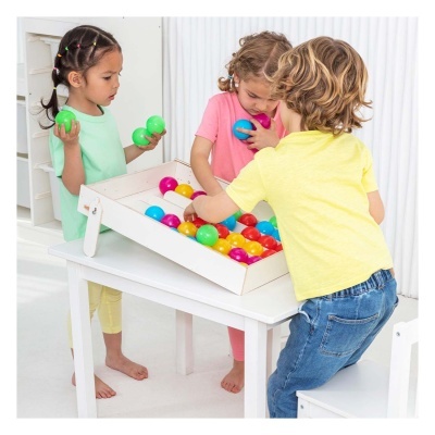 Três crianças a brincar com bolas coloridas numa caixa branca numa mesa branca.