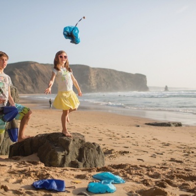 Crianças na praia com roupas coloridas e brinquedos azuis sobre areia perto do mar e rochas