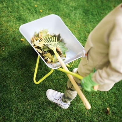 Carrinho de mão branco com estrutura amarela e folhas secas em relva, com pessoa segurando ancinho
