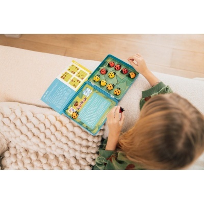 Jogo educativo com peças de joaninhas e patos em caixa azul, folheto e pessoa num sofá claro