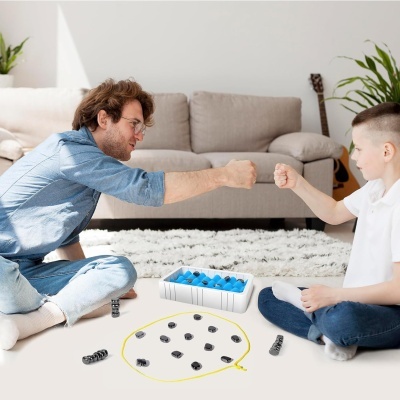 Adulto e criança sentados no chão a jogar jogo de tabuleiro com peças pretas e azuis, caixa branca, círculo amarelo desenhado no chão, sofá cinzento e plantas ao fundo.