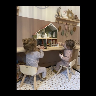 Duas crianças brincando com casa de bonecas em sala decorada com parede rosa e bege