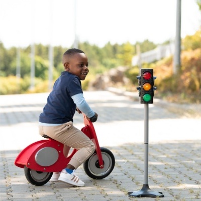Menino numa mota de brinquedo vermelha junto a um semáforo infantil ao ar livre