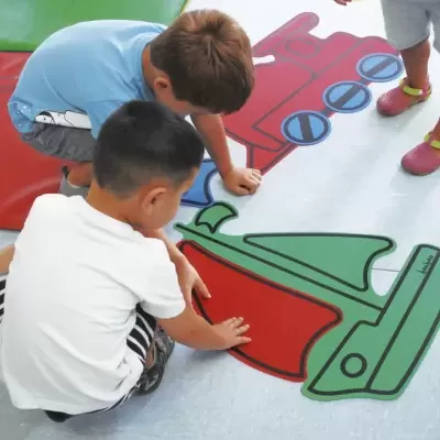 Crianças brincando em tapete colorido com desenhos grandes no chão em sala.