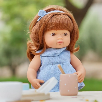 Boneca ruiva com vestido azul e fita na cabeça em mesa com copo cor-de-rosa com paus de gelado