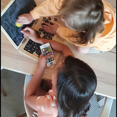 Crianças interagindo com lousa, letras e telemóvel numa mesa de madeira clara