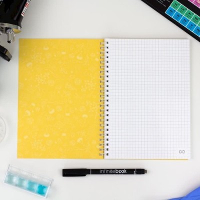 Caderno aberto com páginas quadriculadas, capa amarela e caneta preta 'infinitebook' numa mesa branca.