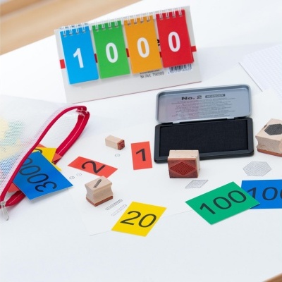 Cartões coloridos com números, carimbos de madeira e almofada de tinta metálica sobre mesa branca.