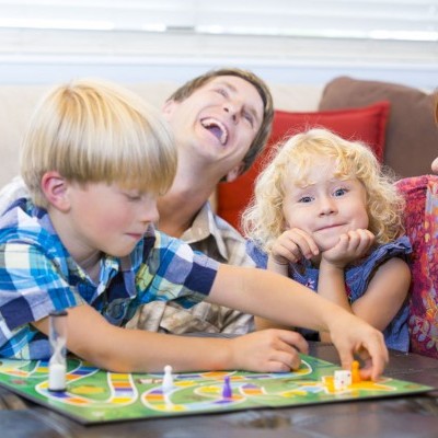 Familia a jogar um jogo de tabuleiro colorido numa sala com sofa azul e almofadas vermelhas