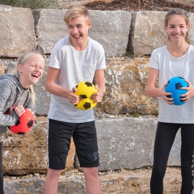 Três crianças com bolas de futebol coloridas posando em ambiente exterior com muros de pedra