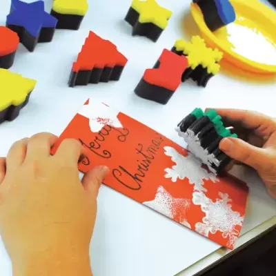 Esponjas de carimbo para atividades de Natal com mãos a usar uma esponja para pintar um cartão vermelho.