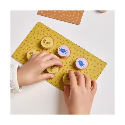 Peças de jogo redondas de madeira com ilustrações de formigas e caranguejos sobre tabuleiro amarelo com padrões alaranjados.