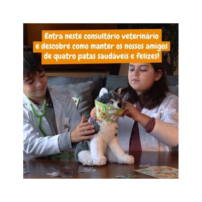 Duas crianças com batas brancas e estetoscópios segurando peluche de cão com cone protetor verde numa sala com mesa de madeira.