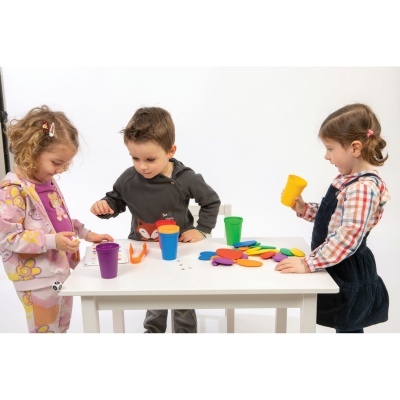 Três crianças a brincar com copos e formas coloridas em mesa branca