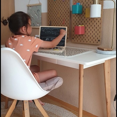 Criança desenhando num tablet sentada à mesa branca com cadeira e painéis de madeira coloridos na parede