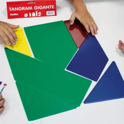 Jogo Tangram Gigante com peças coloridas grandes numa mesa branca e mãos a organizar as peças.