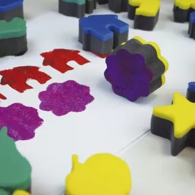 Carimbos de espuma colorida com formas de animais, estrelas e árvores e suas estampas em tinta sobre papel branco.
