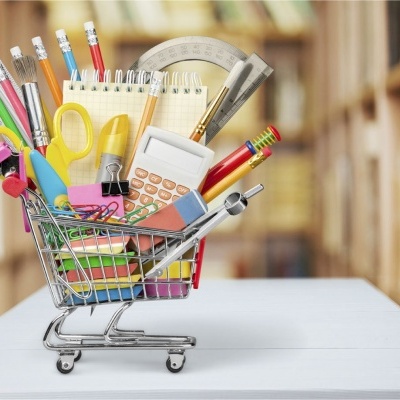 Carrinho de compras miniatura cheio de materiais escolares coloridos numa mesa branca com estantes ao fundo